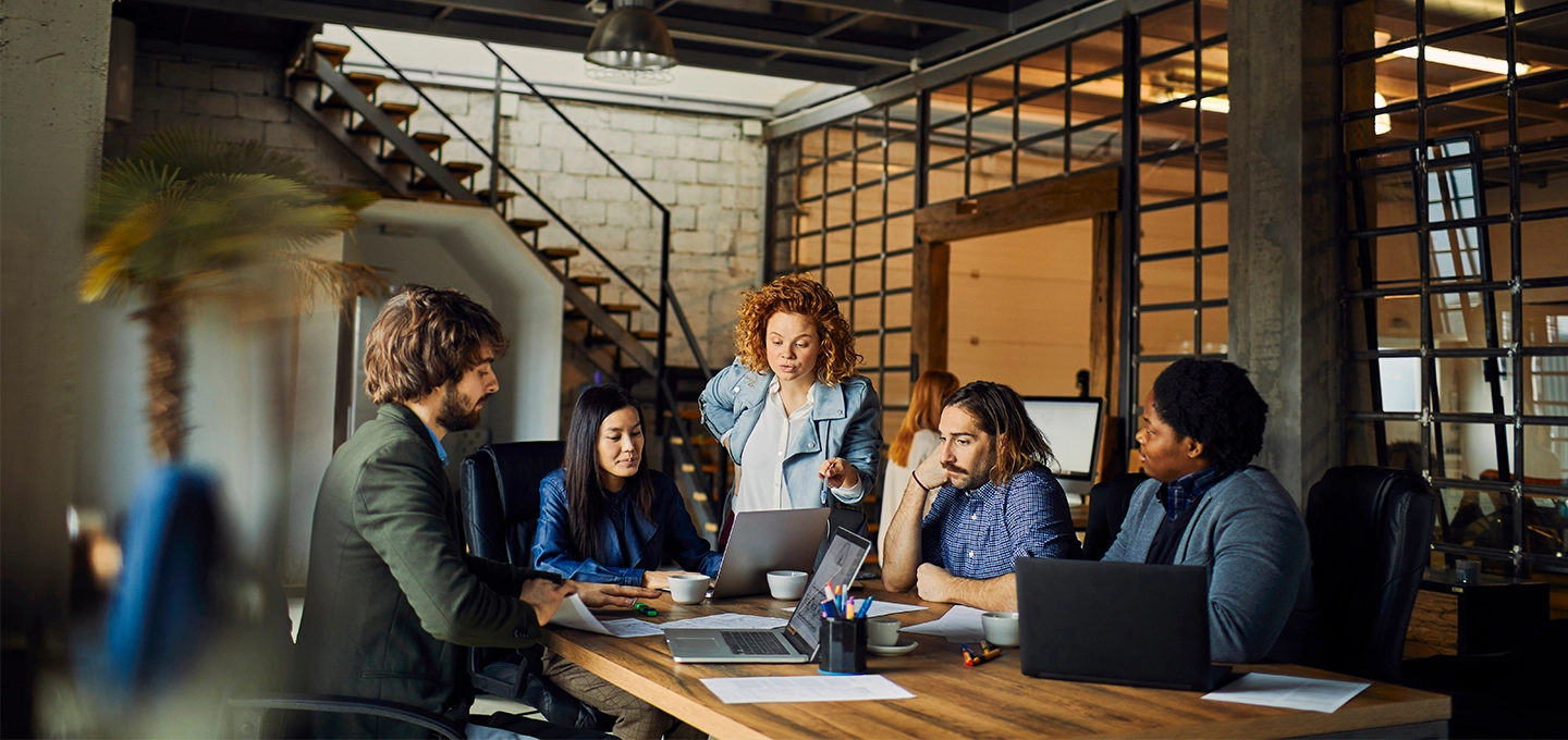 Diverse group of young professionals smiling and collaborating during a creative team meeting in a modern office with laptops and notebooks.